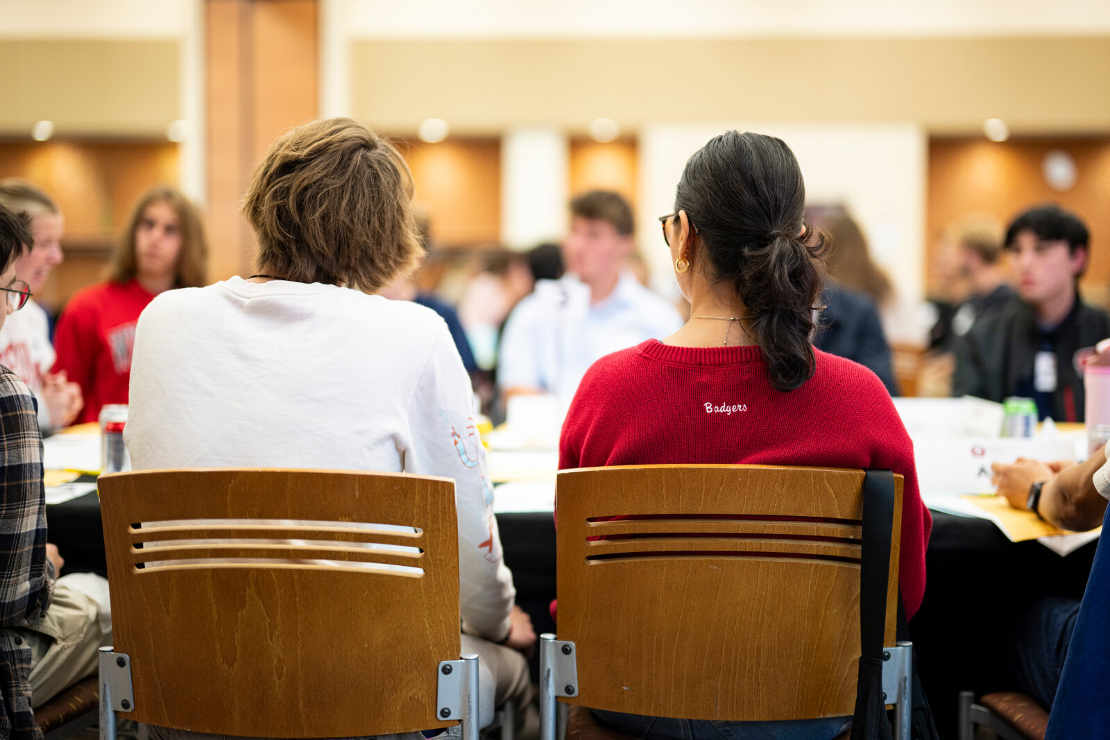 Two people seated in well-lit room, with others blurred in the background.