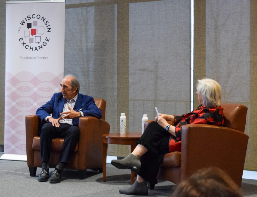 Two people sit in chairs at the head of a room during a discussion.