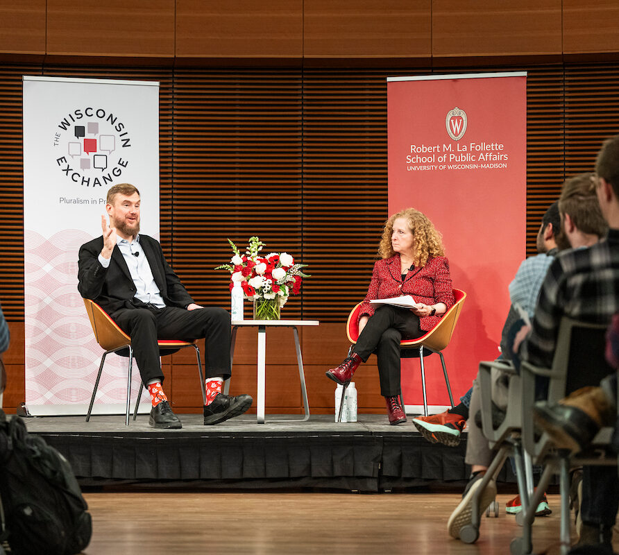 Two people sit onstage in conversation, framed by a Wisconsin Exchange and Robert M. La Follette School of Public Affairs event banner, with an audience watching.