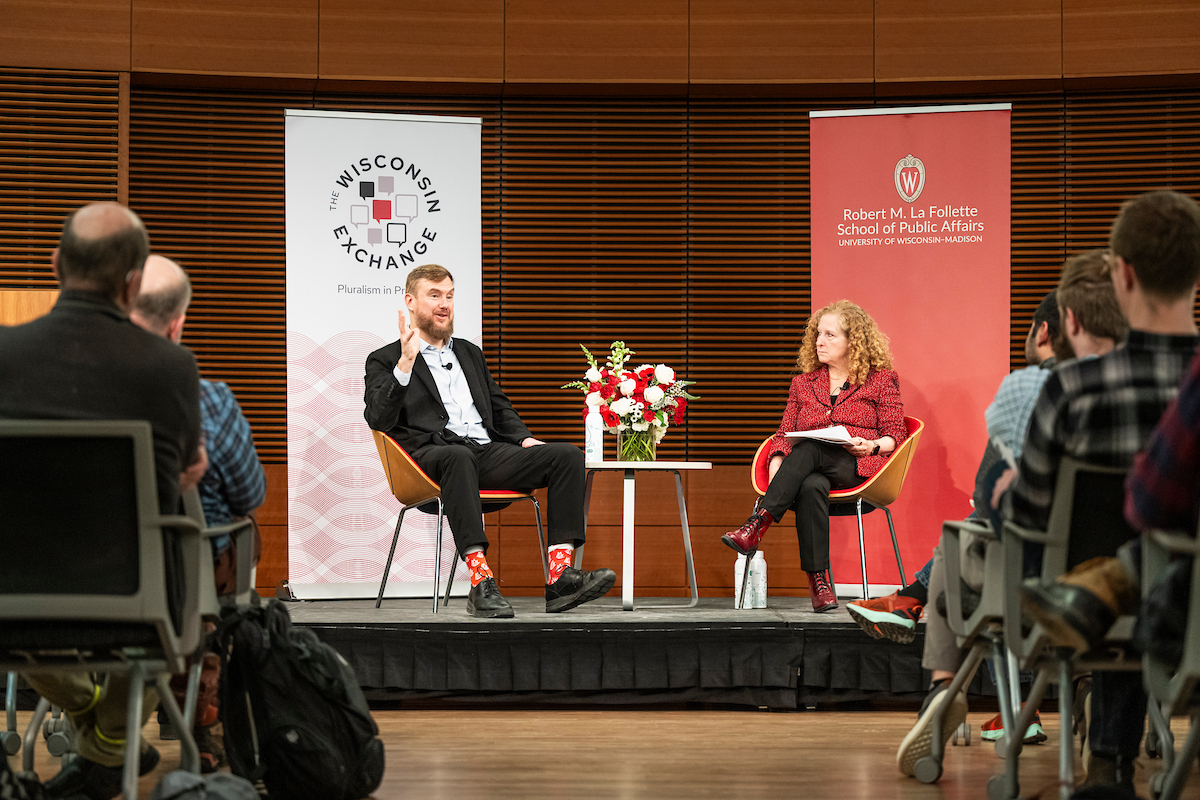 Two people sit onstage in conversation, framed by a Wisconsin Exchange and Robert M. La Follette School of Public Affairs event banner, with an audience watching.