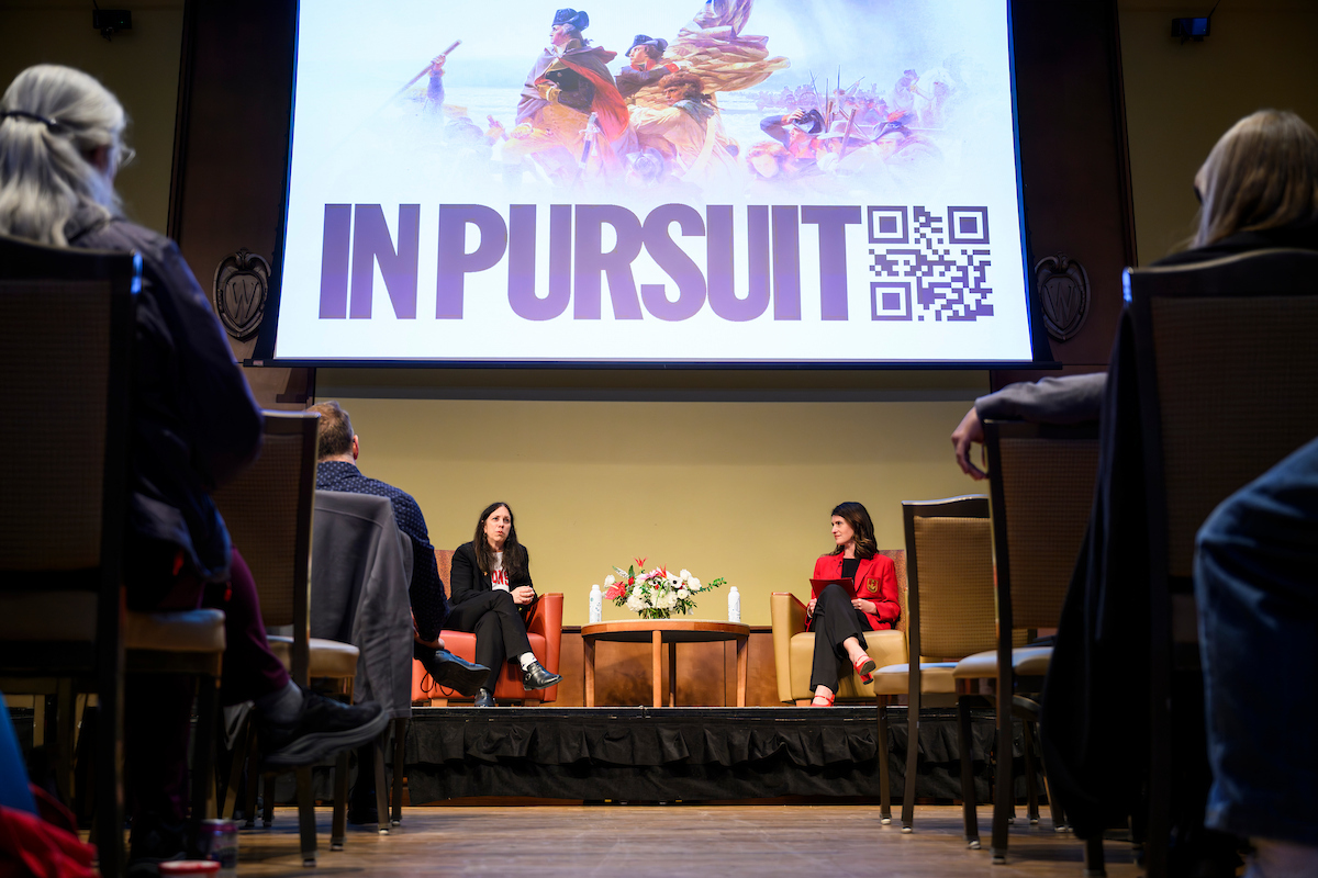 Two people sit onstage in conversation, with large “In Pursuit” text and QR code projected on screen behind them and an audience watching.